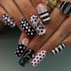 May include: A set of long, square-shaped artificial nails with various designs. Some nails feature black and pink polka dots, others have black and white stripes, and some have silver metal accents and spikes. The nails are a mix of colors and textures.