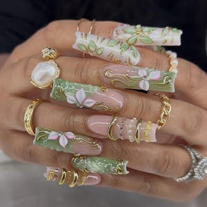 May include: A hand with long, ornate acrylic nails. The nails feature a combination of pink, white, and green designs with gold accents. Some nails have floral embellishments and others have gold chain and ring details. A large pearl ring and other gold rings are also visible.