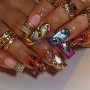 May include: Close-up of a hand with long, decorated nails and gold rings. The nails feature various designs, including tortoiseshell patterns, iridescent accents, and gold embellishments. Some nails have a light pink base, while others are painted in vibrant colors.