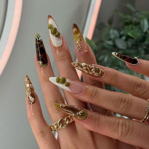 May include: A hand with long, stiletto-shaped acrylic nails. The nails feature various designs, including olive green, brown, white, and gold accents. Some nails have 3D embellishments, such as small green spheres and gold chain details. The nails are a mix of solid colors and patterned designs.