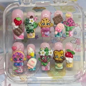 May include: A set of artificial nails in a clear case. Each nail is painted pink and decorated with colorful, cartoonish 3D embellishments, including strawberries, chocolate, and character figures. The designs are whimsical and playful.