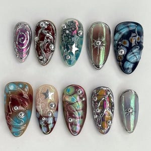 May include: A collection of ten almond-shaped artificial nails. Each nail showcases unique designs with silver metallic accents and embellishments, including swirls, stars, and circular details. The colour palette includes shades of pink, blue, green, and brown.