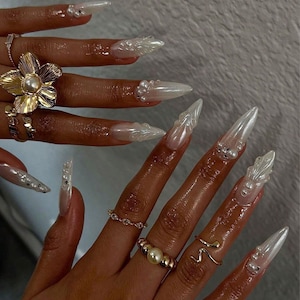May include: Close-up of a hand with long, pointed, pearl-white acrylic nails. The nails are adorned with pearl accents and sculpted, translucent designs. Gold rings, including a flower and a pearl ring, are visible on the fingers.