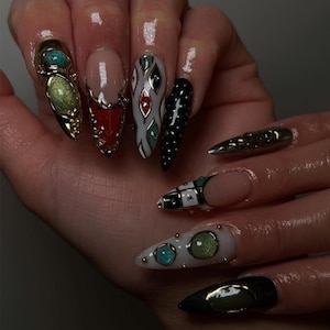 May include: A hand displaying a set of long, pointed, and elaborately decorated artificial nails. The nails feature a variety of designs, including gold embellishments, gemstones, and intricate patterns in colors like red, black, white, and green.