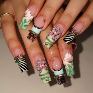 May include: Long square-shaped artificial nails featuring a variety of intricate designs including 3D pink floral accents, black lace patterns, black and white zebra stripes, and small metallic rings adorned with pink rhinestones on a light green base.