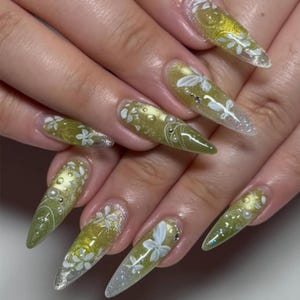 May include: Long, pointed fingernails with a gradient design. The nails transition from a light, shimmery silver at the tips to a golden-green hue at the base. White floral designs and small pearls adorn the nails, creating an elegant look.