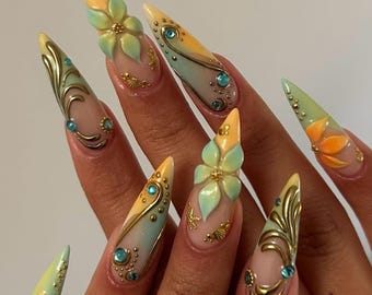 May include: Stiletto-shaped nails featuring a gradient of soft yellow and mint green. The design includes three-dimensional sculpted flowers, metallic gold swirling filigree patterns, small gold beads, and embedded light blue rhinestones.