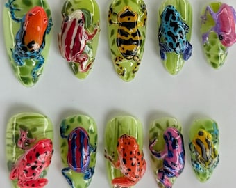 May include: A set of ten artificial nails, each featuring a unique, colorful frog design. The frogs are painted in vibrant hues of red, orange, blue, yellow, and purple, set against a green leaf-shaped background. The nails are oval-shaped.
