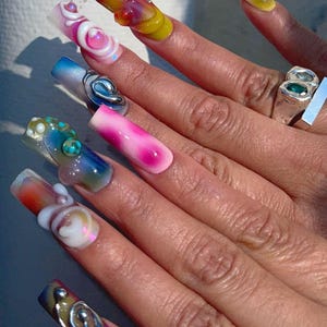 May include: A close-up of a hand with long, colorful acrylic nails. The nails feature various designs, including swirls, gradients, and 3D embellishments. Colors include pink, blue, yellow, and iridescent accents. A silver ring with a green stone is also visible.