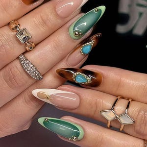 May include: A close-up of manicured nails featuring various designs. Some nails have a tortoiseshell pattern with turquoise accents, while others showcase a French tip with a nude base and gold embellishments. Rings with gemstones adorn the fingers.