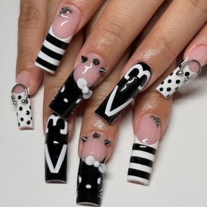 Op de afbeelding: Een set lange, vierkante acrylnagels met diverse ontwerpen. Sommige nagels hebben zwart-witte strepen, stippen of een zwarte doodskistvorm met een wit ontwerp. Andere nagels hebben een roze basis met zilveren spikes en witte accenten.