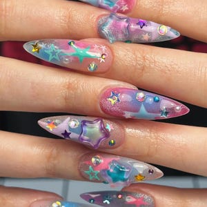 May include: Stiletto-shaped nails featuring a gradient of pink, blue, and purple hues with a glittery finish. The nails are decorated with various three-dimensional star-shaped charms in multiple colors, small iridescent rhinestones, and tiny pearls.