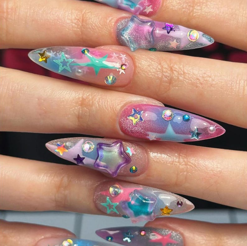 May include: Stiletto-shaped nails featuring a gradient of pink, blue, and purple hues with a glittery finish. The nails are decorated with various three-dimensional star-shaped charms in multiple colors, small iridescent rhinestones, and tiny pearls.
