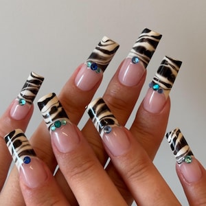 May include: Long square-shaped artificial nails featuring a French manicure base with black and white zebra print tips. Each nail is accented with small, round, multi-colored blue and green rhinestones placed at the base of the patterned tips.