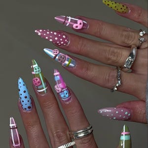 Op de afbeelding: Een hand met lange, puntige acrylnagels. De nagels hebben verschillende ontwerpen, waaronder stippen, strepen en 3D-versieringen zoals harten en knopen. Kleuren zijn onder meer roze, blauw, groen en geel.