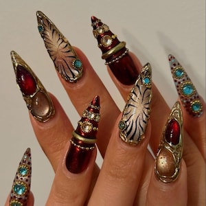 May include: Stiletto-shaped artificial nails featuring intricate 3D designs. The set includes deep red metallic finishes, gold-toned filigree accents, embedded clear and turquoise rhinestones, and black-on-cream radial patterns on a pointed nail structure.