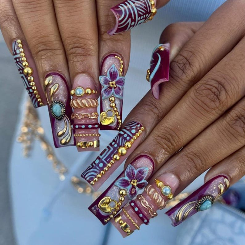 May include: Long, ornate, and embellished artificial nails. The nails feature a combination of burgundy, gold, and light blue colors. Designs include floral accents, beaded details, and intricate patterns. Some nails have a turquoise stone.