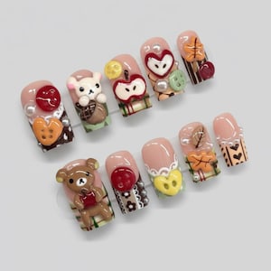 May include: A set of ten artificial nails with a light pink base, each adorned with 3D designs. The designs include a teddy bear, an apple, a heart, and fall leaves, with accents of pearls and buttons.