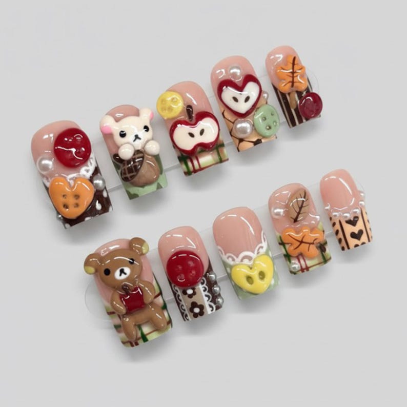 May include: A set of ten artificial nails with a light pink base, each adorned with 3D designs. The designs include a teddy bear, an apple, a heart, and fall leaves, with accents of pearls and buttons.