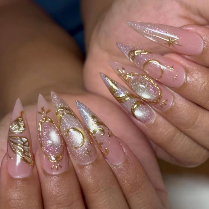 May include: Long, pointed fingernails with a light pink base and glitter accents. Gold embellishments include moons, stars, and ornate filigree designs. The nails have a glamorous, celestial theme.