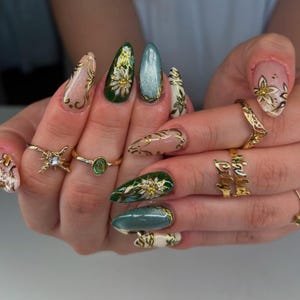 May include: Close-up of manicured nails featuring diverse designs. Nails are painted in green, beige, and white, decorated with gold floral and swirl patterns. Several gold rings with detailed designs are also visible.