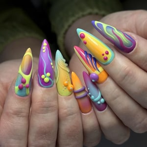 Peut inclure: Gros plan sur une main présentant une manucure avec des ongles en acrylique longs et pointus. Chaque ongle arbore un design unique aux couleurs vives : jaune, orange, violet et bleu, avec des embellissements 3D et des motifs tourbillonnants.
