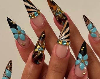 May include: Stiletto-shaped artificial nails featuring a mix of designs including black and tan zebra stripes, leopard print, and tortoise shell patterns. The nails are decorated with three-dimensional light blue flowers and gold rhinestones.