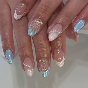 May include: A set of manicured nails with a mermaid theme. The nails feature a variety of designs, including iridescent blue and white shell patterns, pearl accents, and gold starfish embellishments. Some nails have a French tip design with white and iridescent accents.
