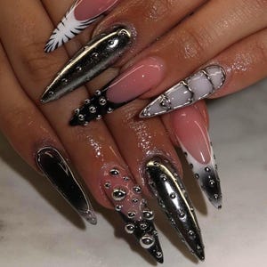 Può includere: Primo piano di unghie acriliche lunghe e appuntite in vari design. Alcune unghie sono cromate, altre nere con borchie argentate, e alcune hanno una french manicure bianca o rosa. Le unghie sono decorate con abbellimenti argentati.