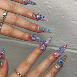 May include: Stiletto-shaped artificial nails featuring a translucent pink and purple gradient base. The nails are decorated with various holographic star-shaped sequins, small metallic charms, and tiny clear rhinestones embedded within the acrylic surface.