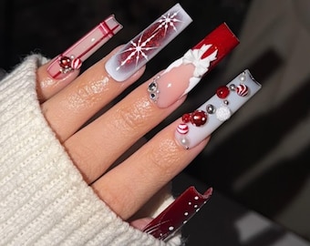 May include: A hand with long, square-shaped, festive Christmas-themed nail art. Designs include snowflakes, plaid, and red and white accents with decorative embellishments. The nails are painted in a variety of colors, including red, white, and clear.