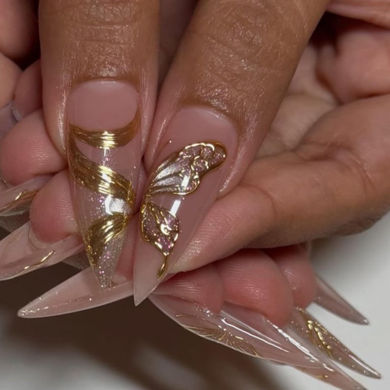 May include: Close-up of long, pointed acrylic nails. The nails are a nude color with gold and glitter accents. One nail features a gold wire design, while another has a gold butterfly. The other nails have gold accents.