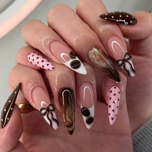 May include: A set of long, almond-shaped artificial nails. The nails feature various designs, including brown polka dots, coffee bean accents, bow details, and a marbled effect. The base color is a natural pink.