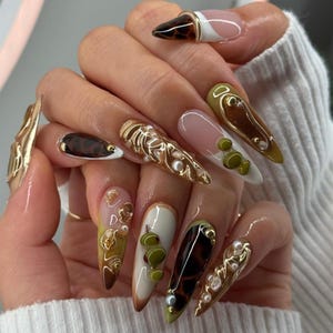 May include: A hand displaying long, almond-shaped acrylic nails. The nails feature a variety of designs, including gold embellishments, tortoiseshell patterns, and pearl accents. Colors include brown, white, green, and gold, creating a luxurious and artistic manicure.
