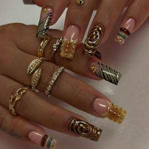 May include: Long square-shaped artificial nails featuring a mix of designs including gold studs, black zebra stripes on a nude base, raised metallic gold swirls, and horizontal bands of green and brown. Several gold rings adorn the fingers.