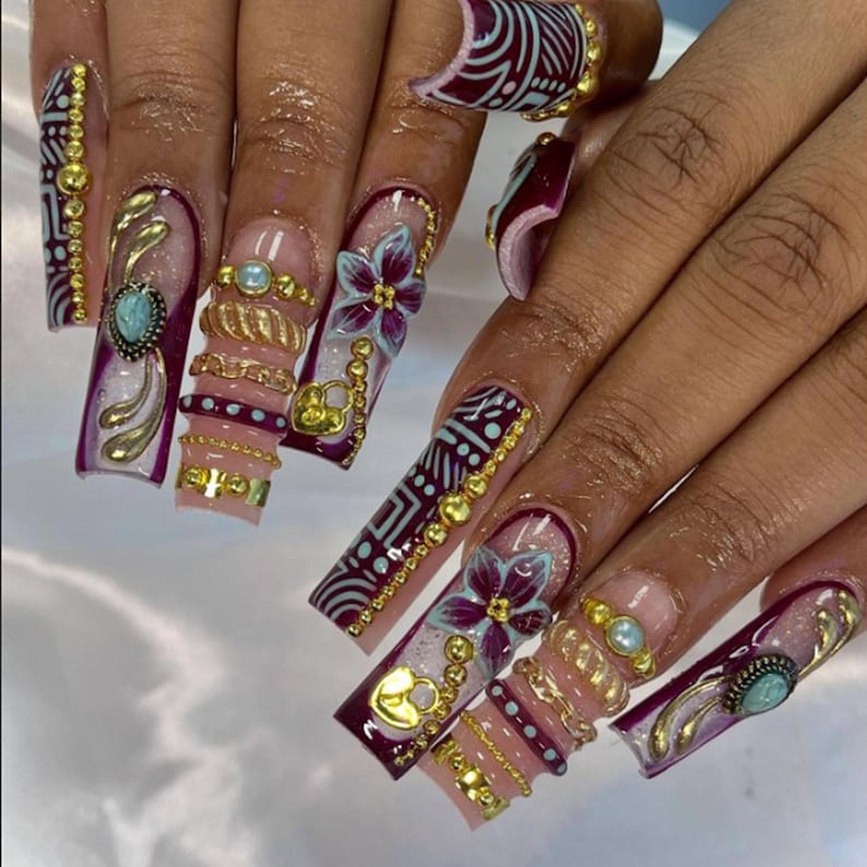 May include: Long, square-shaped artificial nails with intricate designs. The nails feature a base of light pink with burgundy and gold accents. Embellishments include gold chains, beads, and floral designs, with a turquoise stone on some nails.