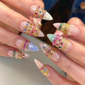 May include: Close-up of manicured nails with a variety of designs. Each nail features a different design, including colorful gems, gold chains, and painted tips. The nails are long and pointed, with a clear base coat.