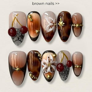 May include: A set of ten brown nail designs. The nails feature various designs, including cherries, floral accents, and gold embellishments. The text "brown nails >>" is at the top of the image.