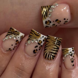 May include: Close-up of a hand with manicured nails. The nails feature a combination of tiger stripe and leopard print designs in gold, black, and white. Rhinestones are placed along the edges of the nails for added detail.