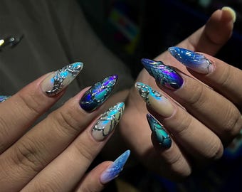 May include: Close-up of a hand with long, pointed, iridescent acrylic nails. The nails feature a gradient of blues, purples, and greens, with silver embellishments. The design resembles a mermaid or dragon scale theme.