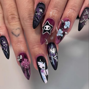 May include: Long almond-shaped nails featuring a dark purple and black gradient base. Decorations include a white skull inside a black star, a painted character face, silver metal charms, and small three-dimensional purple and blue flowers.