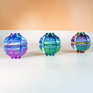 K&ouml;nnte beinhalten: Drei kugelf&ouml;rmige Fidget-Spielzeuge mit einer gitterartigen, segmentierten Oberfl&auml;chenstruktur. Die linke Kugel hat einen blau-violetten Farbverlauf, die mittlere Kugel ist gr&uuml;n-blau und die rechte Kugel hat ein buntes, irisierendes Finish.