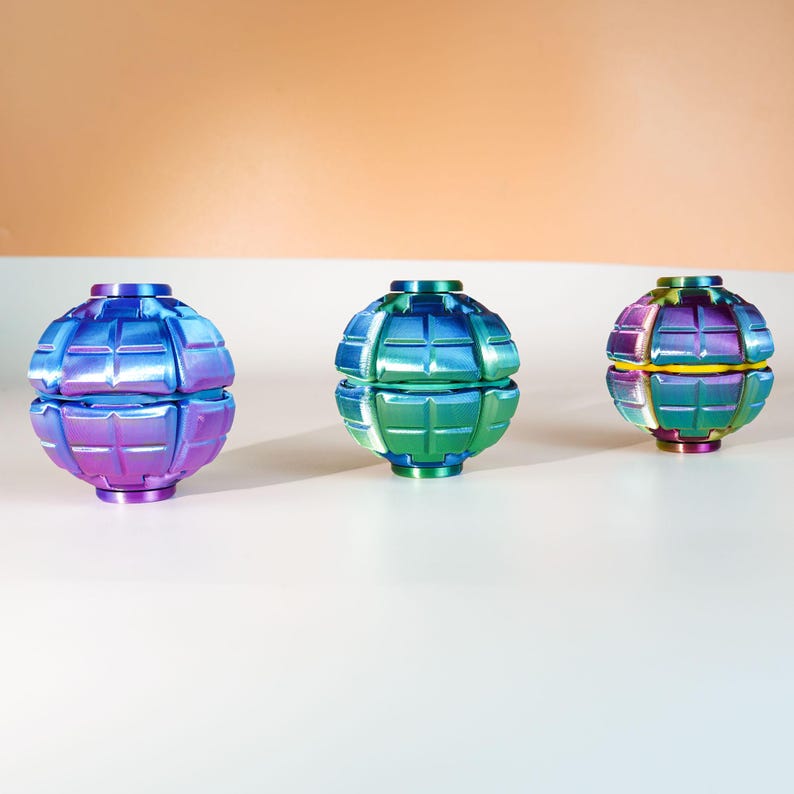 K&ouml;nnte beinhalten: Drei kugelf&ouml;rmige Fidget-Spielzeuge mit einer gitterartigen, segmentierten Oberfl&auml;chenstruktur. Die linke Kugel hat einen blau-violetten Farbverlauf, die mittlere Kugel ist gr&uuml;n-blau und die rechte Kugel hat ein buntes, irisierendes Finish.