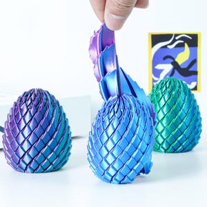 Pode incluir: Três esculturas decorativas de ovos de dragão iridescentes. Uma está sendo parcialmente aberta, revelando uma estrutura em camadas em forma de leque. Os ovos são em tons de azul, roxo e verde. Os ovos têm um padrão geométrico em forma de escamas.