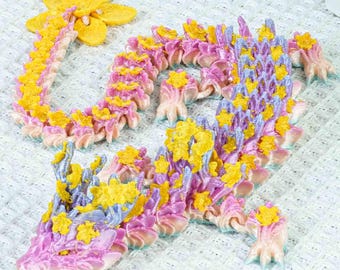 May include: A pink and purple dragon-like creature with yellow flower accents. The creature is made of fabric and has a textured, scaly appearance.