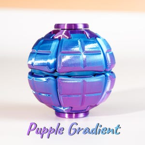K&ouml;nnte beinhalten: Ein kugelf&ouml;rmiges Fidget-Spielzeug mit metallischem Finish, das zwischen Blau- und Violettt&ouml;nen wechselt. Die Oberfl&auml;che zeigt ein Gitter aus erhabenen rechteckigen Segmenten. Unter dem Objekt ist der Text "Purple Gradient" aufgedruckt.