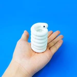 Puede incluir: Un objeto cilíndrico blanco en espiral con un acabado mate liso y un pequeño punto negro cerca de la parte superior. La estructura consta de varios anillos redondeados apilados que forman una forma hueca similar a un resorte.
