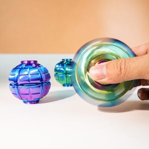 Könnte beinhalten: Zwei irisierende Metall-Fidget-Spielzeuge mit einem segmentierten, kugelförmigen Design in Blau- und Violetttönen. Eines ist unbeweglich, während das andere in Bewegung eingefangen wurde und einen verschwommenen, kreisförmigen Farbverlauf zeigt.