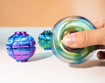 Könnte beinhalten: Zwei kugelförmige Fidget-Spinner mit metallisch irisierender Oberfläche in Blau-, Lila- und Grüntönen. Die Objekte weisen eine segmentierte, gitterartige Oberflächenstruktur auf. Ein Spinner wird in Bewegung gehalten und erzeugt einen verschwommenen Kreiseffekt.