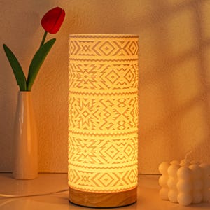 May include: A cylindrical table lamp with a geometric pattern, emitting a warm glow. The lamp has a wooden base and is paired with a white vase holding a red tulip. A white candle is also visible.